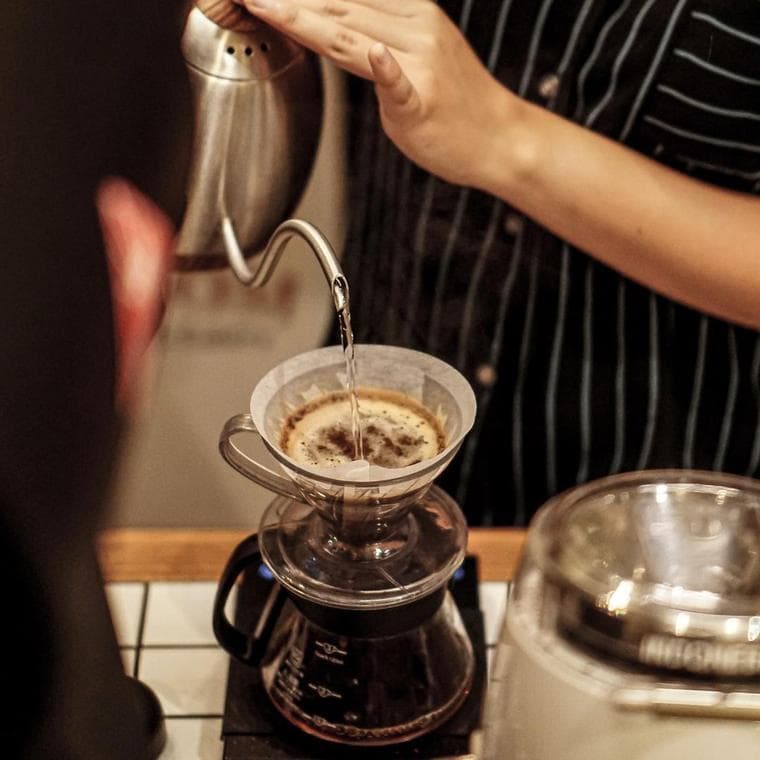 A gooseneck kettle pouring water over a paper filter into a glass pour-over cone